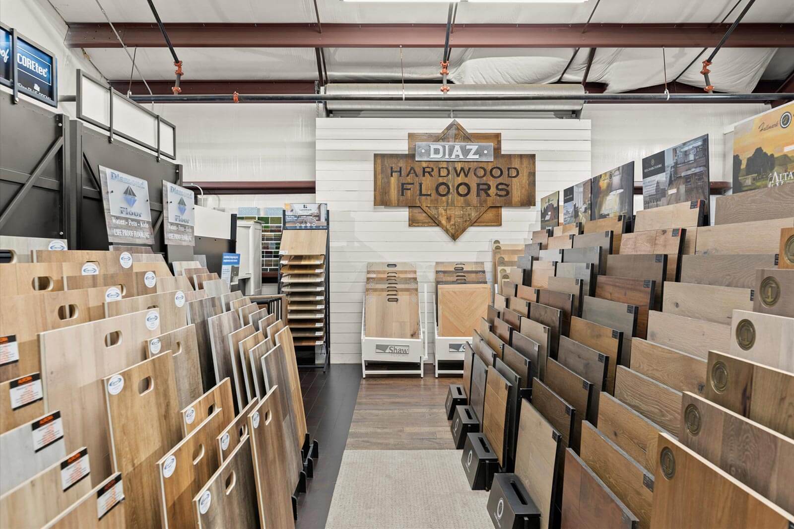 In the center of an aisle containing racks of hardwood floor samples, facing a wooden sign labeled "Diaz Hardwood Floors"