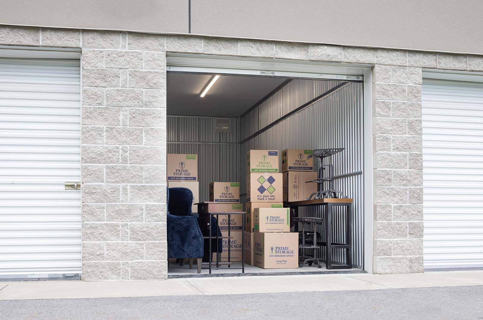 An outside storage facility focused on a single storage unit with the door open, revealing several Prime Storage-branded cardboard boxes
